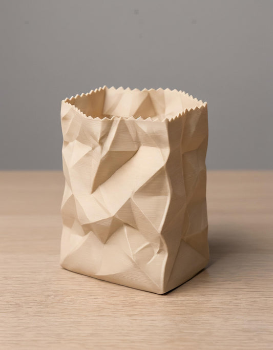 Ein 3D-gedruckter Stiftehalter in cremefarbener PLA-Ausführung mit realistischer zerknüllter Papiertüten-Optik steht auf einer Holzoberfläche. Das geometrische Design zeigt dreidimensionale Falten und Knicke, die einer zusammengeknüllten Papertüte täuschend ähnlich sehen. Der Behälter hat eine gezackte obere Öffnung und eine quadratische Grundform. Perfekt als desk organizer und moderne Büro-Dekoration - ein origineller pen holder für jeden Schreibtisch.
