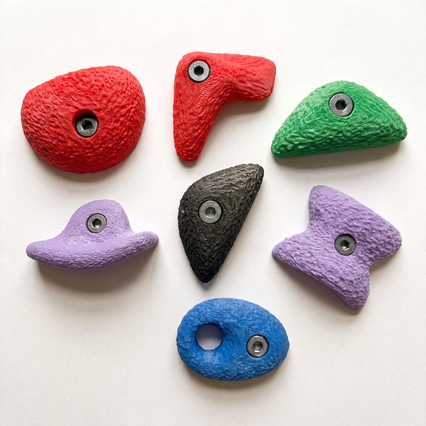 Alle sieben großen Bouldergriff-Magnete im Farbmix, locker angeordnet auf hellem Grund — Kühlschrankmagnete in echten Klettergriff-Formen, handgefertigt im 3D-Druck.