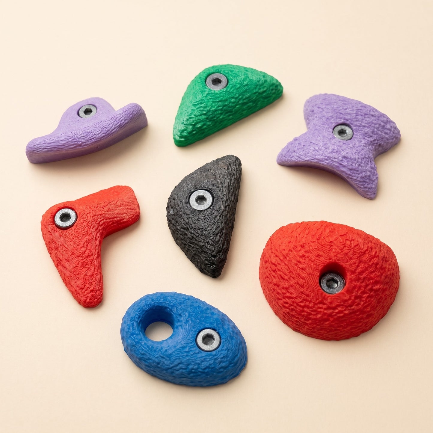 Alle sieben großen Bouldergriff-Magnete im Farbmix auf warmem Cremehintergrund — Klettergriff-Formen Sloper, Crimp, Bowtie, Wave, Pinch, Pocket und Jug als Kühlschrankmagnete.