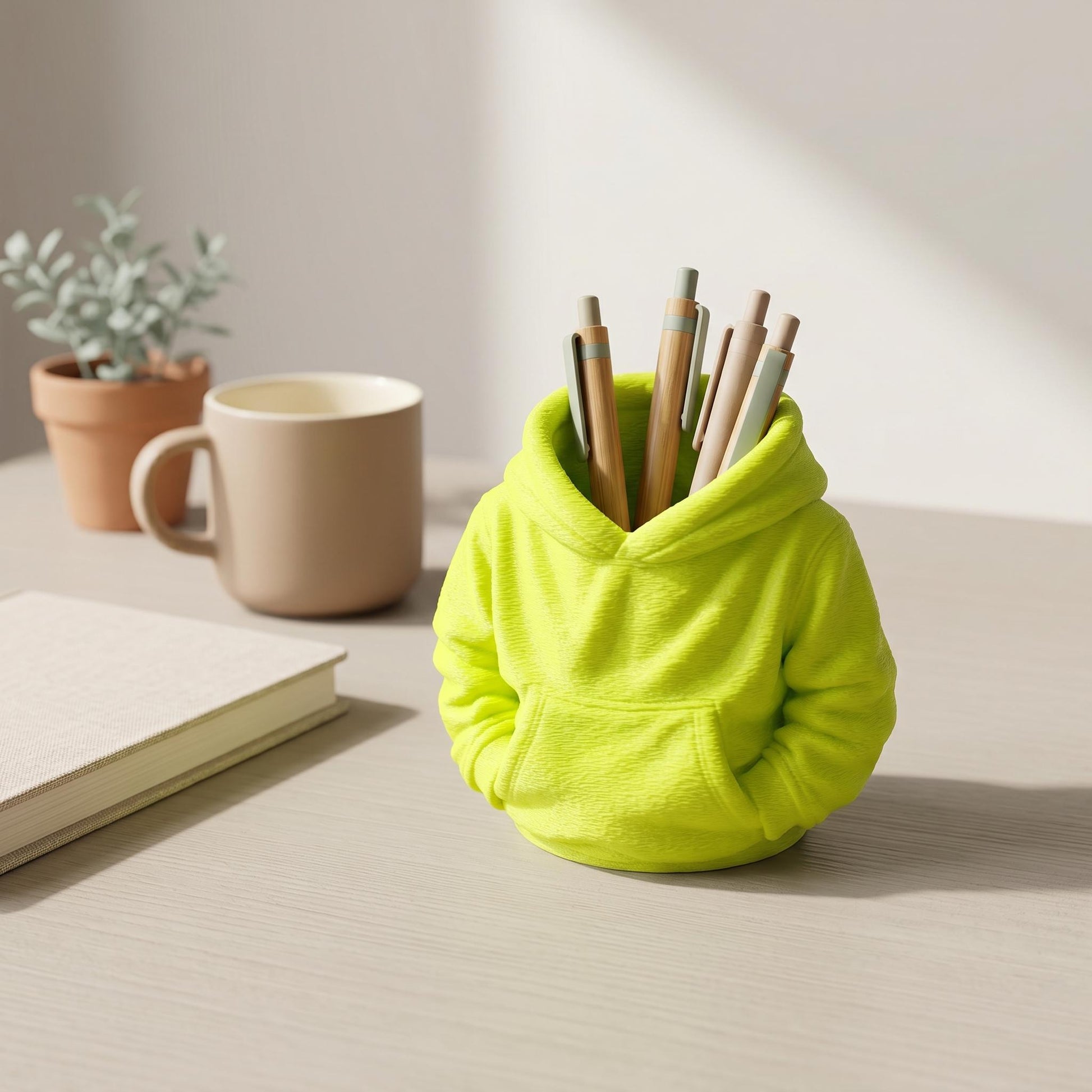Niedlicher 3D-gedruckter Stiftehalter in leuchtend grünem Hoodie-Design auf hellem Holzschreibtisch. Der kawaii Pen Holder aus PLA-Material fasst mehrere Stifte in der Kapuze und fungiert als praktischer Schreibtisch Organizer. Perfekt als Abschiedsgeschenk Lehrerin oder Erzieherin, ideal für Büro-Deko. Maße 11×10×7 cm, ca. 90g leicht. Daneben stehen Kaffeetasse, Notizbuch und Pflanze im Tontopf.