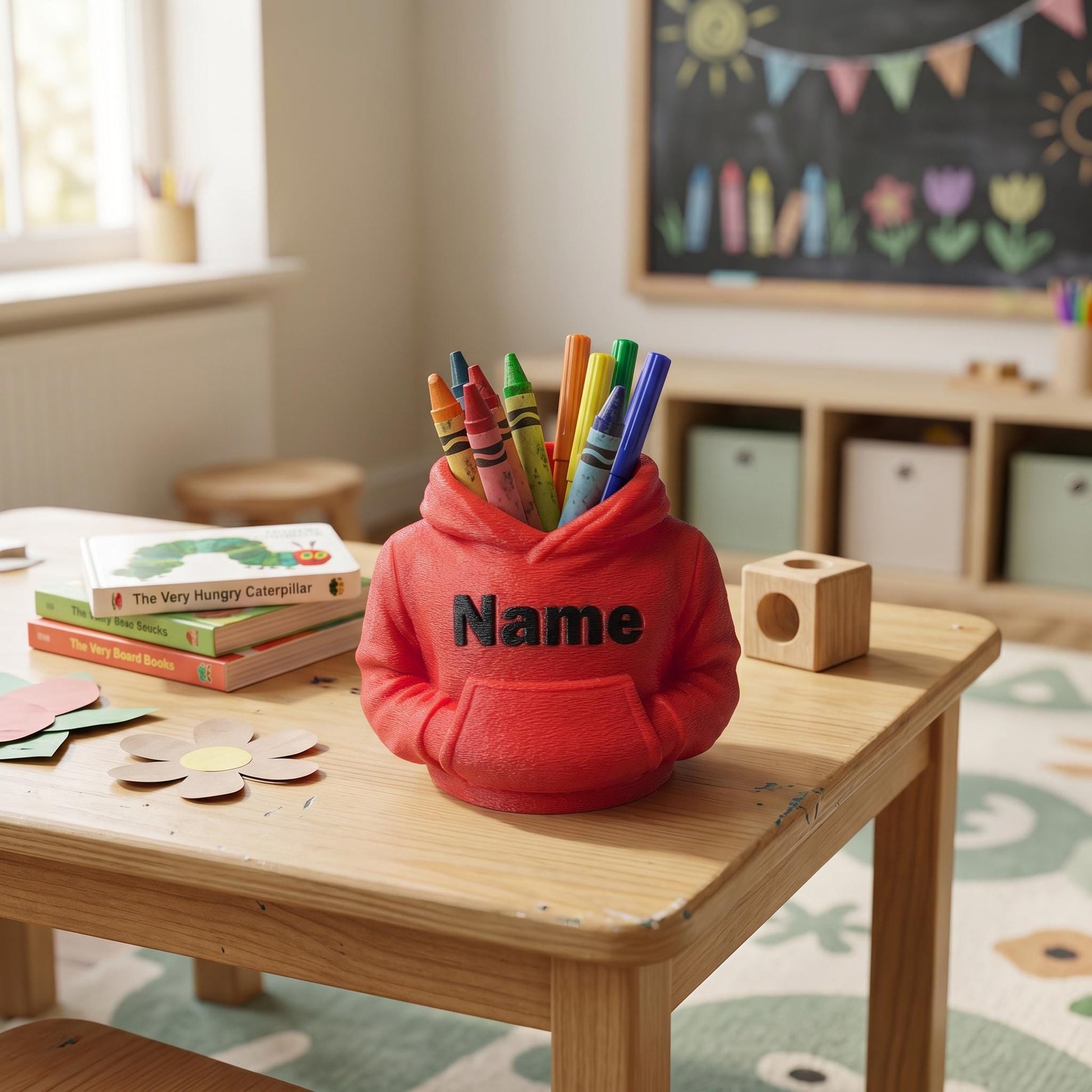 3D-gedruckter Stiftehalter in roter Hoodie-Form mit personalisiertem Namen auf hellbraunem Holztisch. Der PLA-Organizer hält bunte Buntstifte und Marker. Perfekt als Abschiedsgeschenk für Lehrerin oder Erzieherin, Schulgeschenk zur Einschulung oder Schultüte Inhalt. Maße 11x10x7cm. Im Hintergrund Kinderbücher und Bastelutensilien in hellem Kinderzimmer mit Tafel und bunter Wimpelkette.