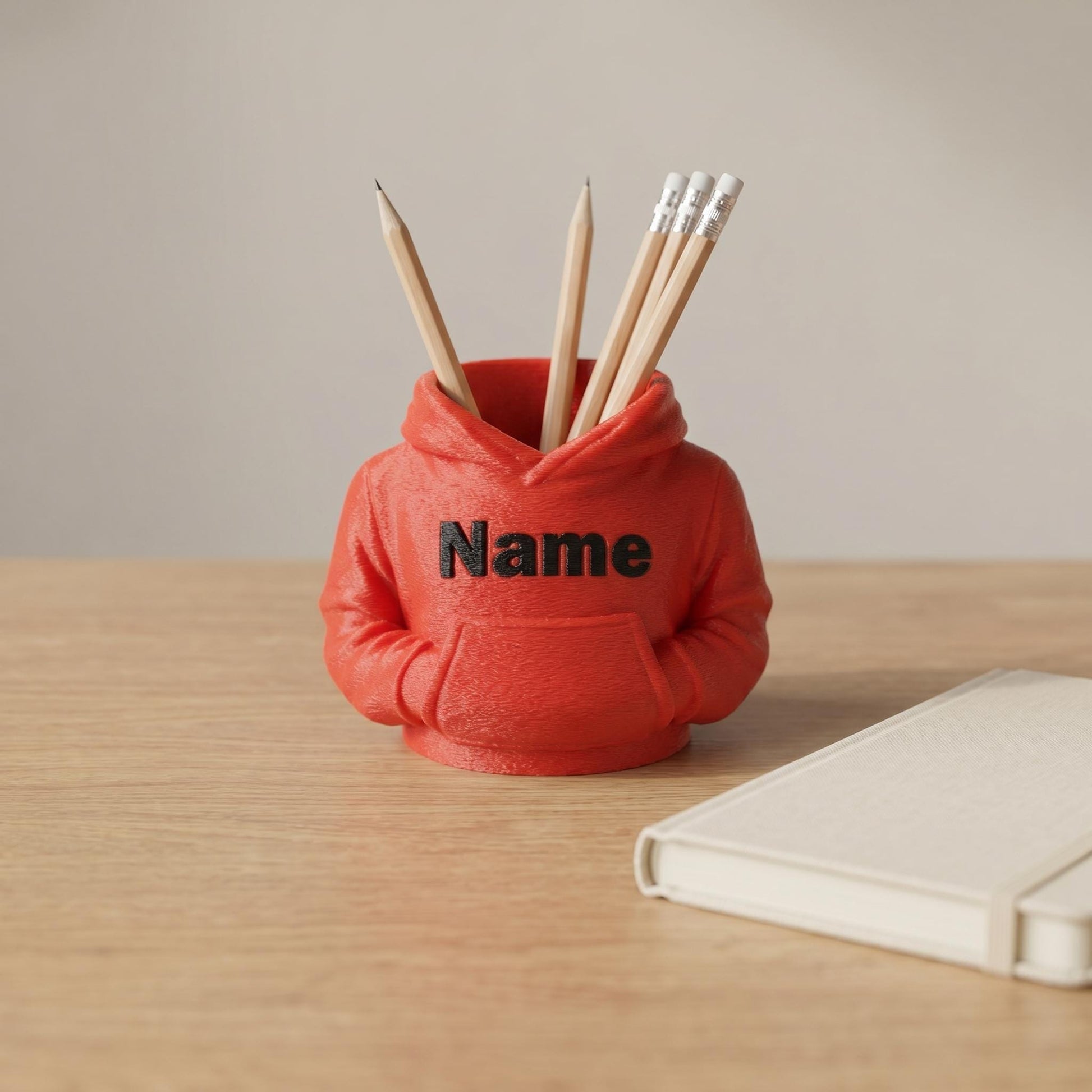 3D-gedruckter Stiftehalter in Form eines roten Hoodies mit Kapuze und Bauchtasche. Der Schreibtisch-Organizer aus rotem PLA-Material zeigt den schwarzen Schriftzug Name auf der Vorderseite und enthält mehrere Holzstifte und Bleistifte mit Radiergummi. Das funktionale Desk-Organizer-Design eignet sich perfekt als Abschiedsgeschenk für Lehrerin oder Erzieherin, zur Einschulung oder als Schulgeschenk. Steht auf hellem Holzschreibtisch neben einem weißen Notizbuch.