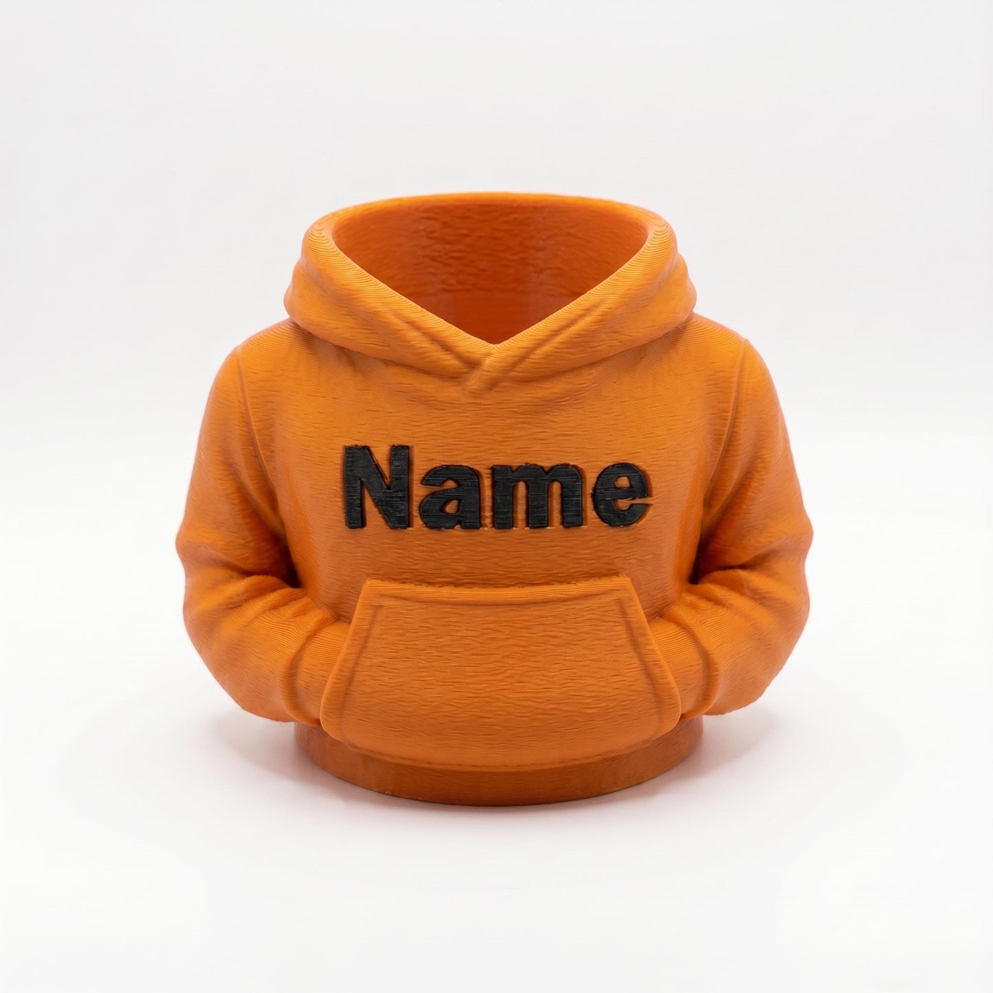 Ein orangefarbener 3D-gedruckter Stiftehalter in Form eines Hoodies mit schwarzer Name-Beschriftung auf der Brust und einer Bauchtasche. Das Design zeigt realistische Hoodie-Details wie Kapuze, Kordelzüge und Ärmelbündchen. Der Schreibtisch-Organizer aus PLA-Material eignet sich perfekt als Abschiedsgeschenk für Lehrerin oder Erzieherin, Schulgeschenk oder Büro-Deko. Personalisierbar mit Wunschname. Maße: 11×10×7 cm.