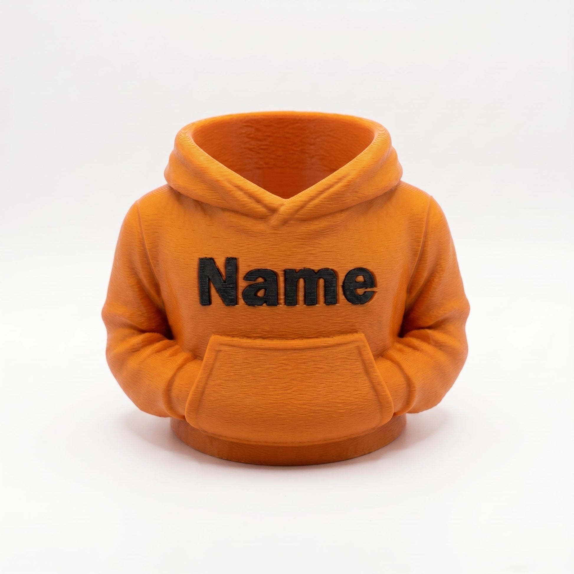 Ein orangefarbener 3D-gedruckter Stiftehalter in Form eines Hoodies mit schwarzer Name-Beschriftung auf der Brust und einer Bauchtasche. Das Design zeigt realistische Hoodie-Details wie Kapuze, Kordelzüge und Ärmelbündchen. Der Schreibtisch-Organizer aus PLA-Material eignet sich perfekt als Abschiedsgeschenk für Lehrerin oder Erzieherin, Schulgeschenk oder Büro-Deko. Personalisierbar mit Wunschname. Maße: 11×10×7 cm.