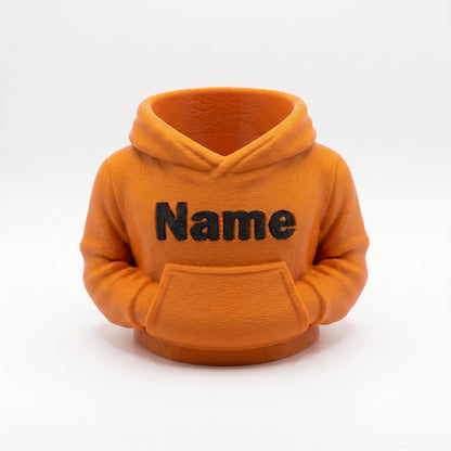 Ein orangefarbener 3D-gedruckter Stiftehalter in Form eines Hoodies mit schwarzer Name-Beschriftung auf der Brust und einer Bauchtasche. Das Design zeigt realistische Hoodie-Details wie Kapuze, Kordelzüge und Ärmelbündchen. Der Schreibtisch-Organizer aus PLA-Material eignet sich perfekt als Abschiedsgeschenk für Lehrerin oder Erzieherin, Schulgeschenk oder Büro-Deko. Personalisierbar mit Wunschname. Maße: 11×10×7 cm.
