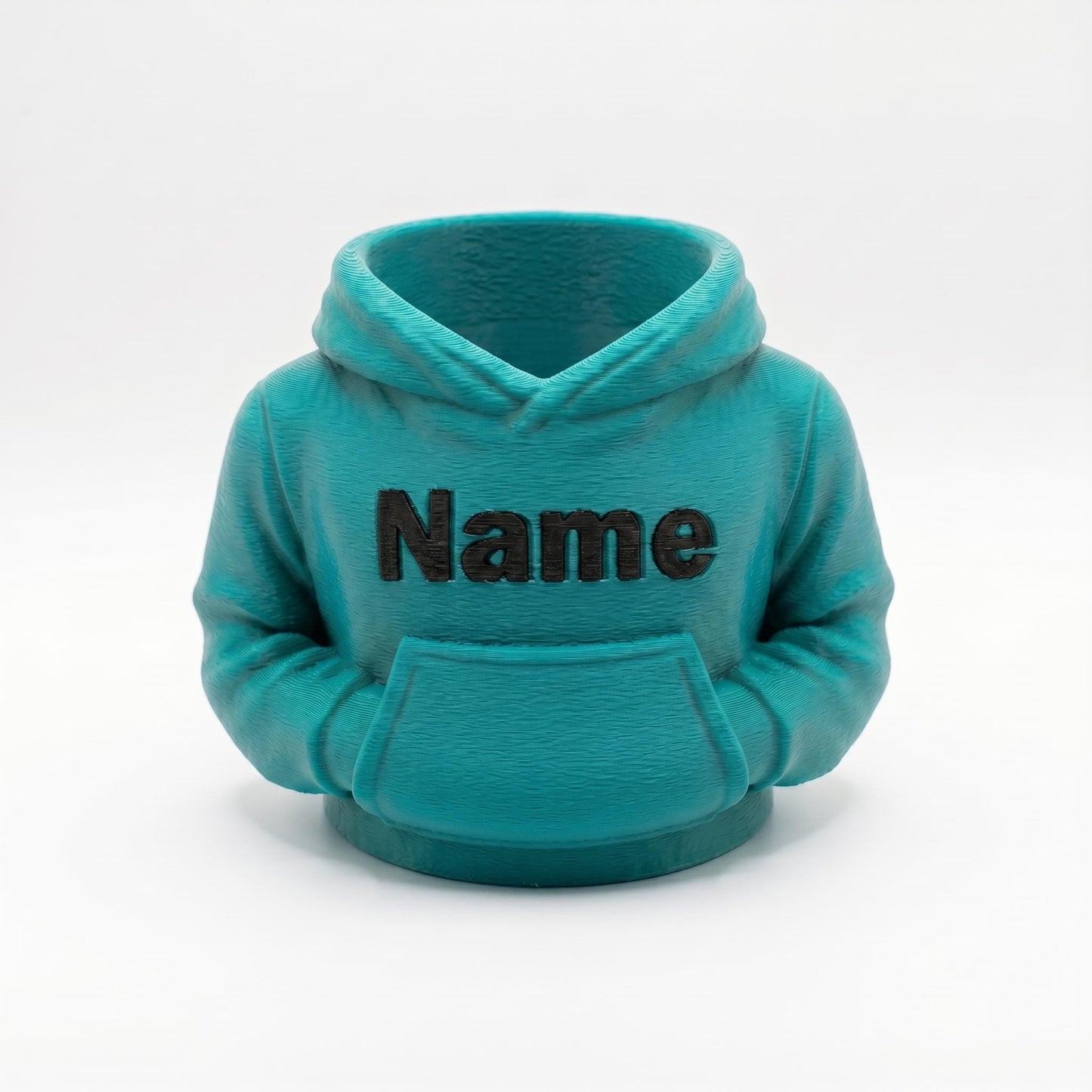 Ein türkisfarbener 3D-gedruckter Stiftehalter in Form eines Miniatur-Hoodies mit der Aufschrift Name auf der Brust. Der detailgetreue Desk Organizer zeigt realistische Hoodie-Strukturen mit Kapuze und Bauchtasche. Perfektes Abschiedsgeschenk für Lehrerin oder Erzieherin, individuell personalisierbar. Maße 11x10x7 cm, aus hochwertigem PLA-Material. Idealer Schreibtisch Organizer für Stifte und kleine Büroartikel. Auch als Schulgeschenk oder Mitgebsel geeignet.