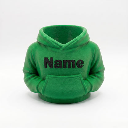 3D-gedruckter Stiftehalter in grüner Hoodie-Form mit schwarzem Name Schriftzug auf der Brust. Der detaillierte Organizer zeigt realistische Kapuze, Kängurutasche und Ärmel mit sichtbaren Drucklinien. Perfekt als Abschiedsgeschenk Lehrerin, Schulgeschenk oder Schreibtisch Organizer. Maße 11×10×7 cm, aus grünem PLA im 3D Druck hergestellt. Ideal für Einschulung oder als personalisierte Büro-Deko.