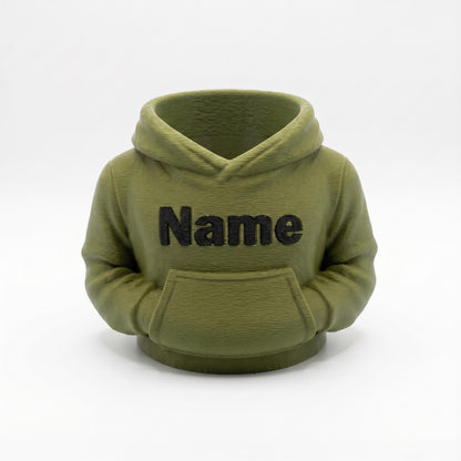 3D-gedruckter Stiftehalter in olivgrüner Farbe im niedlichen Hoodie-Design mit schwarzem Name Schriftzug auf der Vorderseite. Der Organizer verfügt über eine Kapuze und Bauchtasche für Stifte und Büroartikel. Perfekt als Abschiedsgeschenk Lehrerin oder Erzieherin, Schulgeschenk oder Schreibtisch Organizer. Personalisierbar mit Wunschname. Maße: 11x10x7cm, umweltfreundliches PLA Material. Ideal für Büro, Klassenzimmer oder Kinderzimmer.