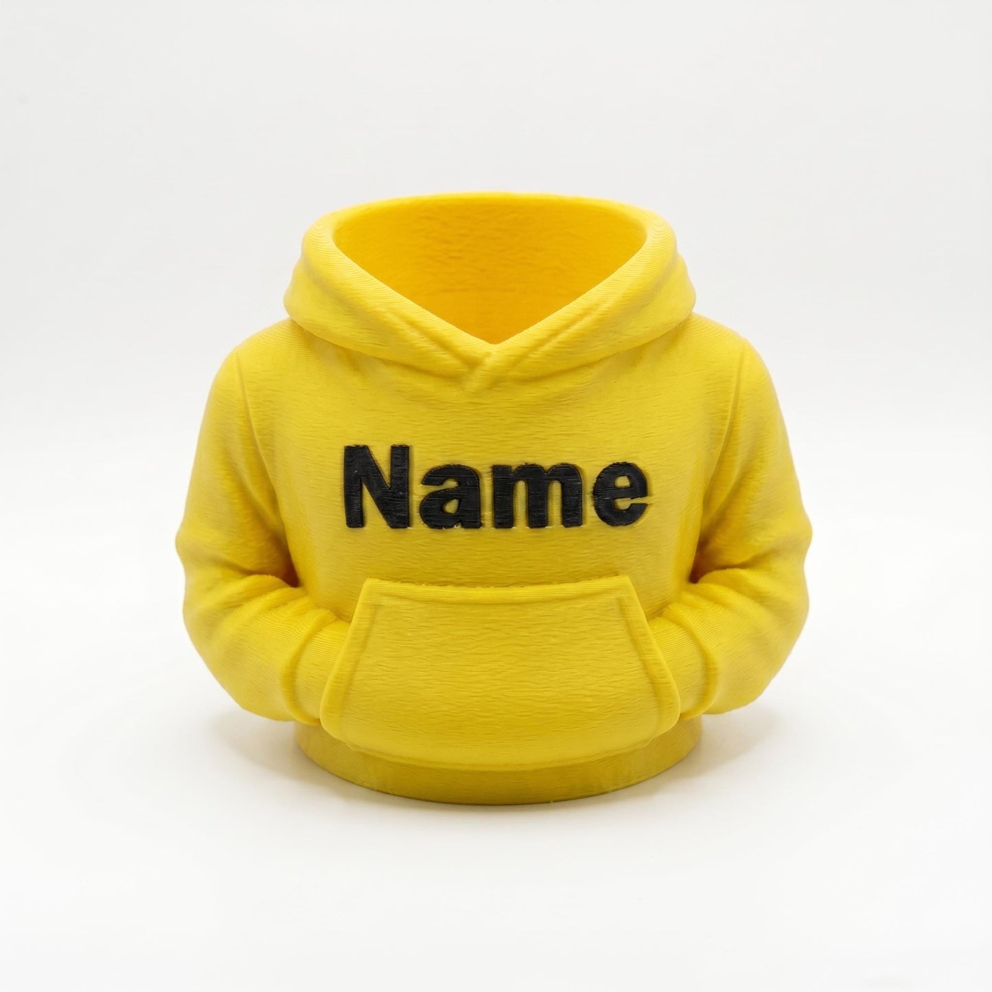Gelber 3D-gedruckter Stiftehalter in Form eines Hoodies mit schwarzem Name-Schriftzug auf der Brust. Der Miniatur-Kapuzenpullover aus PLA-Material verfügt über eine Bauchtasche als Stifthalter und realistische Hoodie-Details wie Kapuze und Ärmel. Perfekter Schreibtisch Organizer als Abschiedsgeschenk Lehrerin oder Erzieherin, Schulgeschenk zur Einschulung oder personalisierte Büro-Deko. Maße: 11×10×7cm, individuell anpassbar.