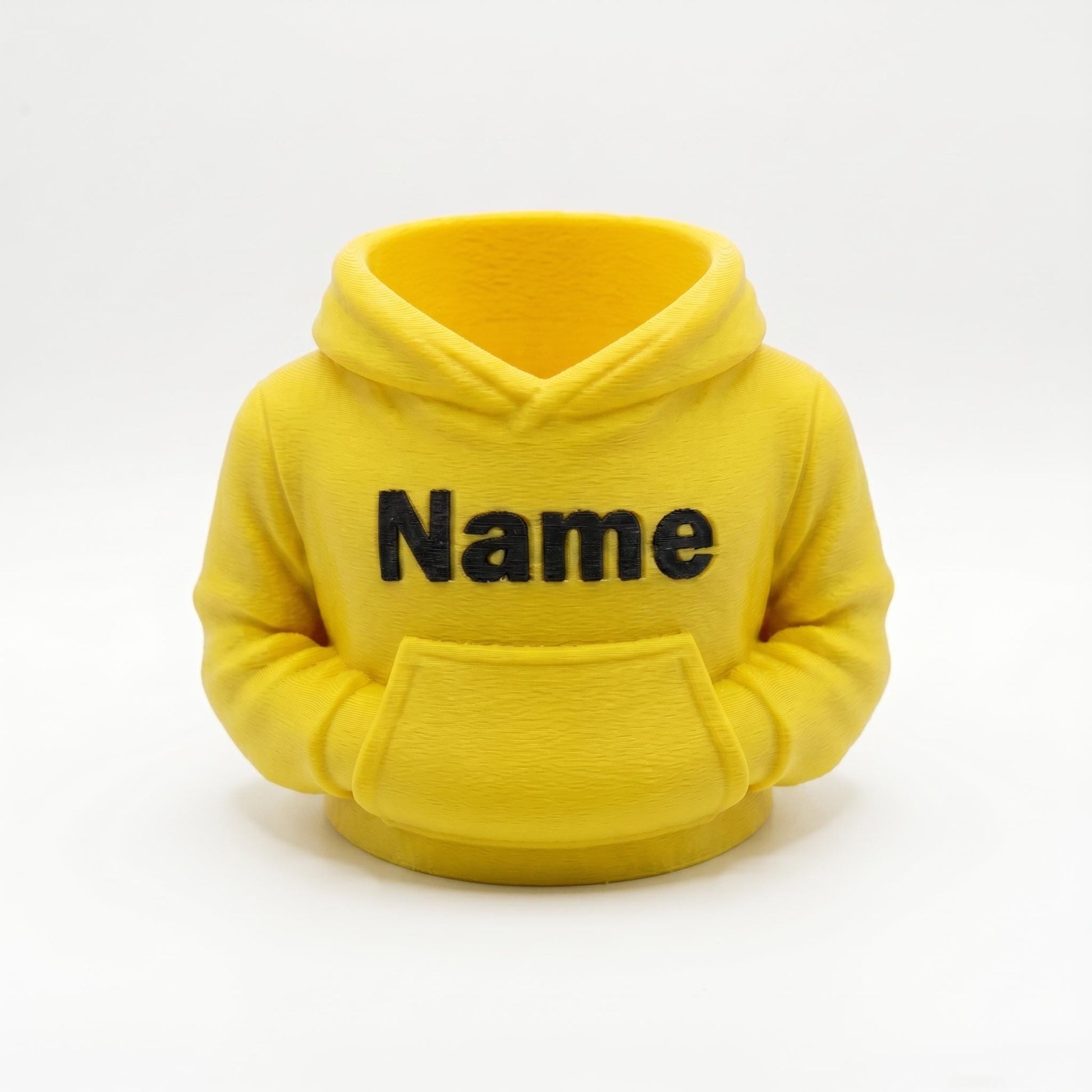 Gelber 3D-gedruckter Stiftehalter in Form eines Hoodies mit schwarzem Name-Schriftzug auf der Brust. Der Miniatur-Kapuzenpullover aus PLA-Material verfügt über eine Bauchtasche als Stifthalter und realistische Hoodie-Details wie Kapuze und Ärmel. Perfekter Schreibtisch Organizer als Abschiedsgeschenk Lehrerin oder Erzieherin, Schulgeschenk zur Einschulung oder personalisierte Büro-Deko. Maße: 11×10×7cm, individuell anpassbar.