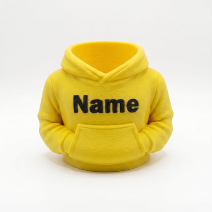 Gelber 3D-gedruckter Stiftehalter in Form eines Hoodies mit schwarzem Name-Schriftzug auf der Brust. Der Miniatur-Kapuzenpullover aus PLA-Material verfügt über eine Bauchtasche als Stifthalter und realistische Hoodie-Details wie Kapuze und Ärmel. Perfekter Schreibtisch Organizer als Abschiedsgeschenk Lehrerin oder Erzieherin, Schulgeschenk zur Einschulung oder personalisierte Büro-Deko. Maße: 11×10×7cm, individuell anpassbar.