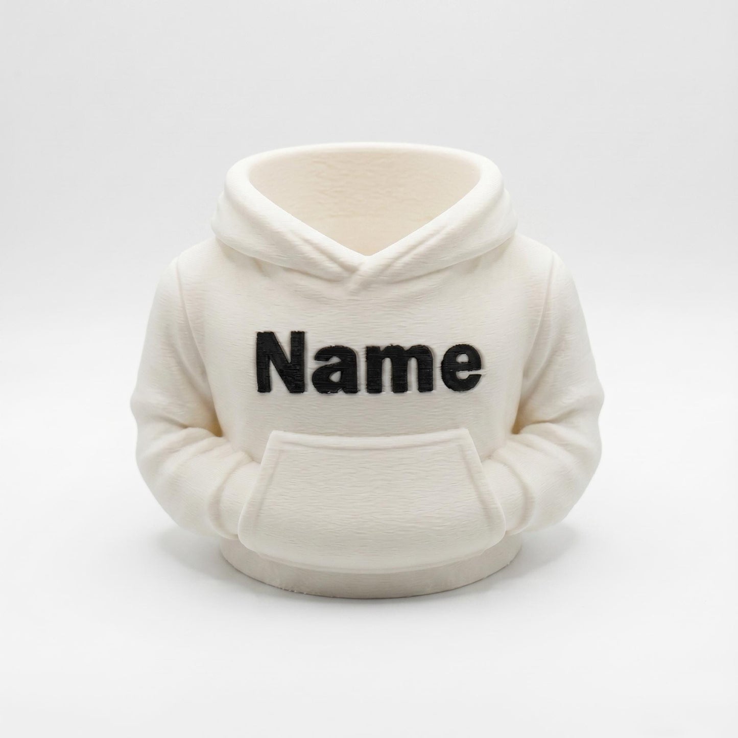 Individueller 3D-gedruckter Stiftehalter in cremefarbener Hoodie-Optik mit schwarzem Name-Aufdruck. Der praktische Desk Organizer aus PLA-Material verfügt über eine Bauchtasche für Stifte und Büromaterial. Perfekt als Abschiedsgeschenk für Lehrerin oder Erzieherin, zur Einschulung oder als Schreibtisch-Deko. Maße 11×10×7cm, personalisierbar mit Wunschname. Moderner 3D Druck für individuelle Büro-Organisation.