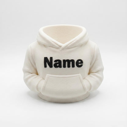 Individueller 3D-gedruckter Stiftehalter in cremefarbener Hoodie-Optik mit schwarzem Name-Aufdruck. Der praktische Desk Organizer aus PLA-Material verfügt über eine Bauchtasche für Stifte und Büromaterial. Perfekt als Abschiedsgeschenk für Lehrerin oder Erzieherin, zur Einschulung oder als Schreibtisch-Deko. Maße 11×10×7cm, personalisierbar mit Wunschname. Moderner 3D Druck für individuelle Büro-Organisation.