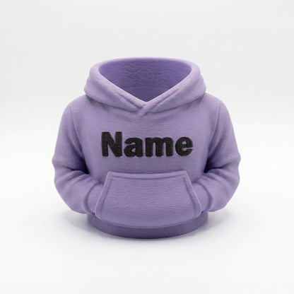 Ein 3D-gedruckter Stiftehalter in Form eines Hoodies in matter Lavendel-Farbe mit schwarzem Name-Schriftzug auf der Vorderseite. Der kompakte Organizer verfügt über eine Kapuze und Kängurutasche, perfekt als Schreibtisch-Deko. Ideales Abschiedsgeschenk für Lehrerin oder Erzieherin, Schulgeschenk oder Mitgebsel. Aus robustem PLA-Material im 3D-Druck hergestellt. Personalisierbar für individuelle Gestaltung.