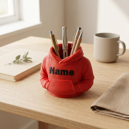Roter 3D-gedruckter Stiftehalter im niedlichen Hoodie-Design mit schwarzem Name-Schriftzug auf der Brust. Der kleine Kapuzenpullover aus PLA-Material steht auf einem hellen Holzschreibtisch und hält verschiedene Stifte in der Kapuze. Perfekter Schreibtisch Organizer als Abschiedsgeschenk Lehrerin oder Erzieherin. Maße: 11x10x7cm. Personalisierbar mit Wunschname. Ideal für Büro-Deko und Schreibtisch-Organisation.