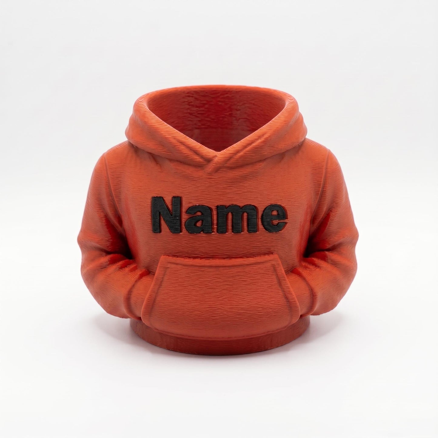 Ein oranger 3D-gedruckter Stiftehalter in Form eines Hoodies mit schwarzem Name Schriftzug auf der Brust. Der Miniatur-Hoodie verfügt über eine Kapuze und Bauchtasche, perfekt als Schreibtisch Organizer für Stifte und Büromaterial. Ideales Abschiedsgeschenk für Lehrerin oder Erzieherin, personalisierbar mit Wunschnamen. Maße 11x10x7cm, aus PLA im 3D Druck hergestellt. Originelle Büro Deko und Schulgeschenk für den Schreibtisch.