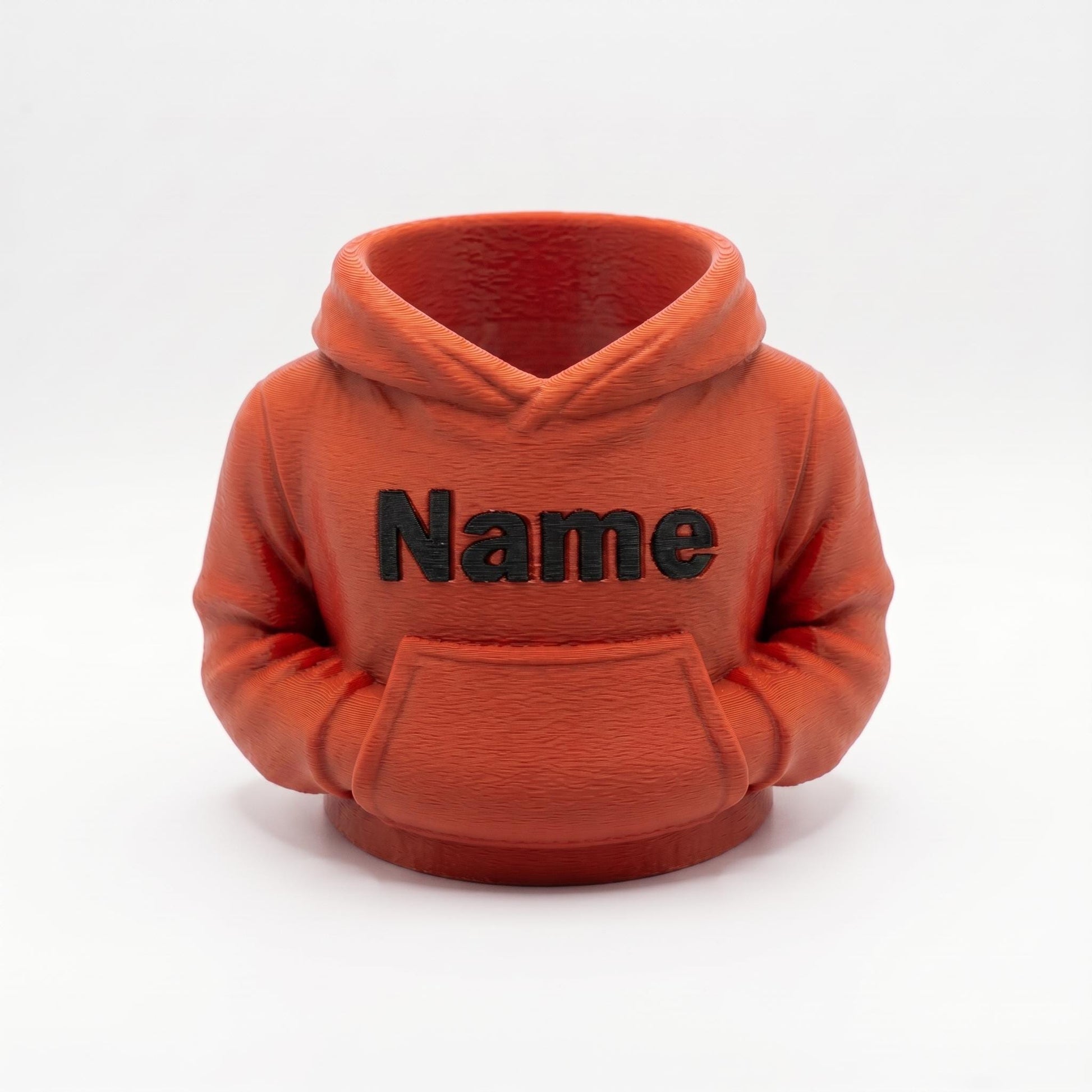 Ein oranger 3D-gedruckter Stiftehalter in Form eines Hoodies mit schwarzem Name Schriftzug auf der Brust. Der Miniatur-Hoodie verfügt über eine Kapuze und Bauchtasche, perfekt als Schreibtisch Organizer für Stifte und Büromaterial. Ideales Abschiedsgeschenk für Lehrerin oder Erzieherin, personalisierbar mit Wunschnamen. Maße 11x10x7cm, aus PLA im 3D Druck hergestellt. Originelle Büro Deko und Schulgeschenk für den Schreibtisch.
