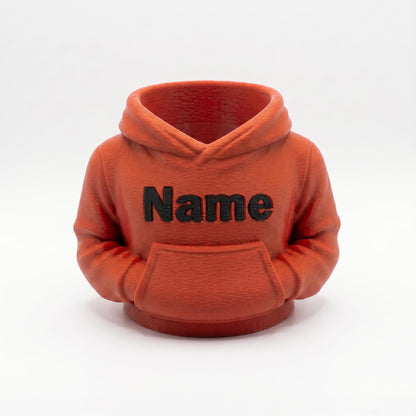 Ein oranger 3D-gedruckter Stiftehalter in Form eines Hoodies mit schwarzem Name Schriftzug auf der Brust. Der Miniatur-Hoodie verfügt über eine Kapuze und Bauchtasche, perfekt als Schreibtisch Organizer für Stifte und Büromaterial. Ideales Abschiedsgeschenk für Lehrerin oder Erzieherin, personalisierbar mit Wunschnamen. Maße 11x10x7cm, aus PLA im 3D Druck hergestellt. Originelle Büro Deko und Schulgeschenk für den Schreibtisch.