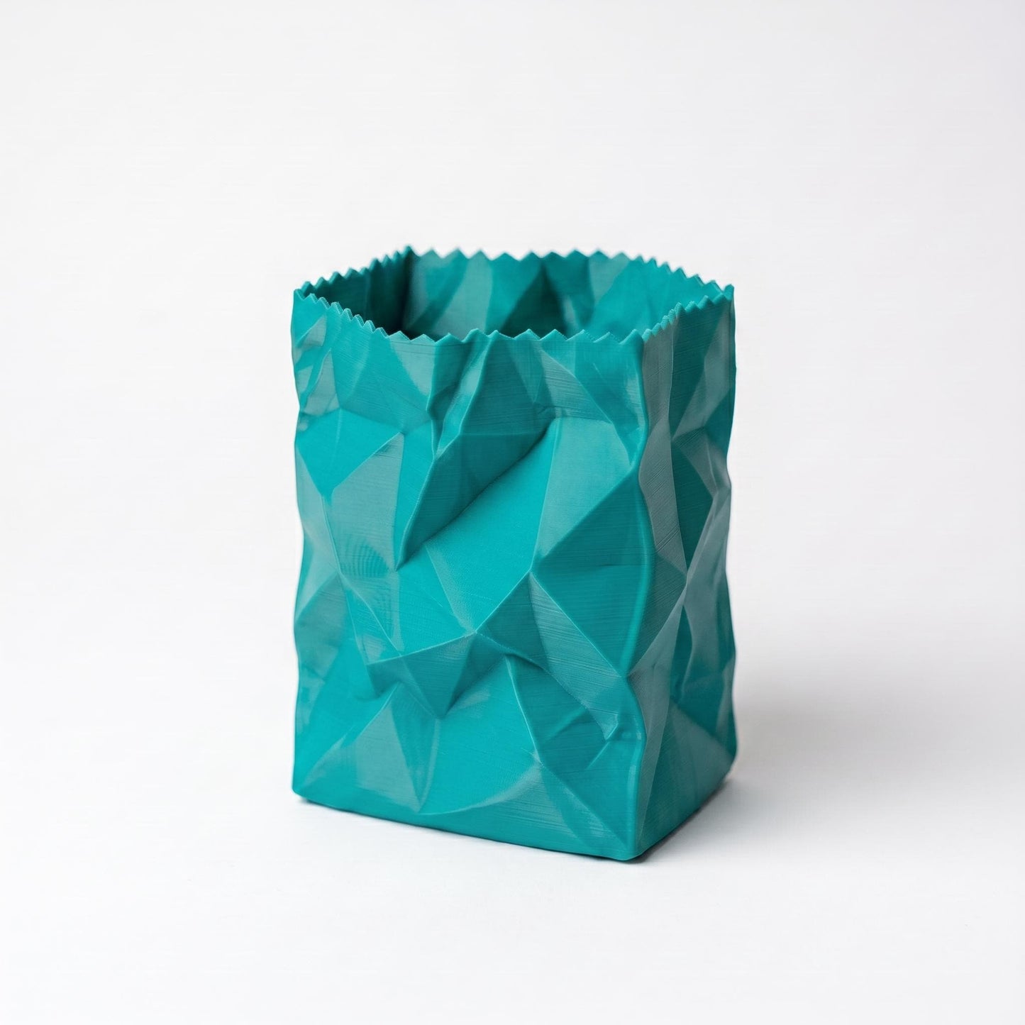 Ein türkisfarbener 3D-gedruckter Stiftehalter im zerknüllten Papiertüten-Design steht auf weißem Hintergrund. Das geometrische Facettenmuster erzeugt ein modernes, skulpturales Aussehen mit scharf definierten Kanten und Flächen. Der desk organizer aus PLA-Filament zeigt eine matte Oberfläche und einen gezackten oberen Rand, perfekt als pen holder für den Schreibtisch oder als originelles Schulgeschenk.