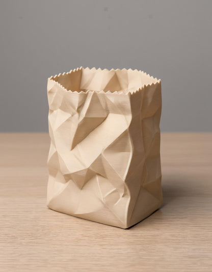 Ein 3D-gedruckter Stiftehalter in cremefarbener PLA-Ausführung mit realistischer zerknüllter Papiertüten-Optik steht auf einer Holzoberfläche. Das geometrische Design zeigt dreidimensionale Falten und Knicke, die einer zusammengeknüllten Papertüte täuschend ähnlich sehen. Der Behälter hat eine gezackte obere Öffnung und eine quadratische Grundform. Perfekt als desk organizer und moderne Büro-Dekoration - ein origineller pen holder für jeden Schreibtisch.