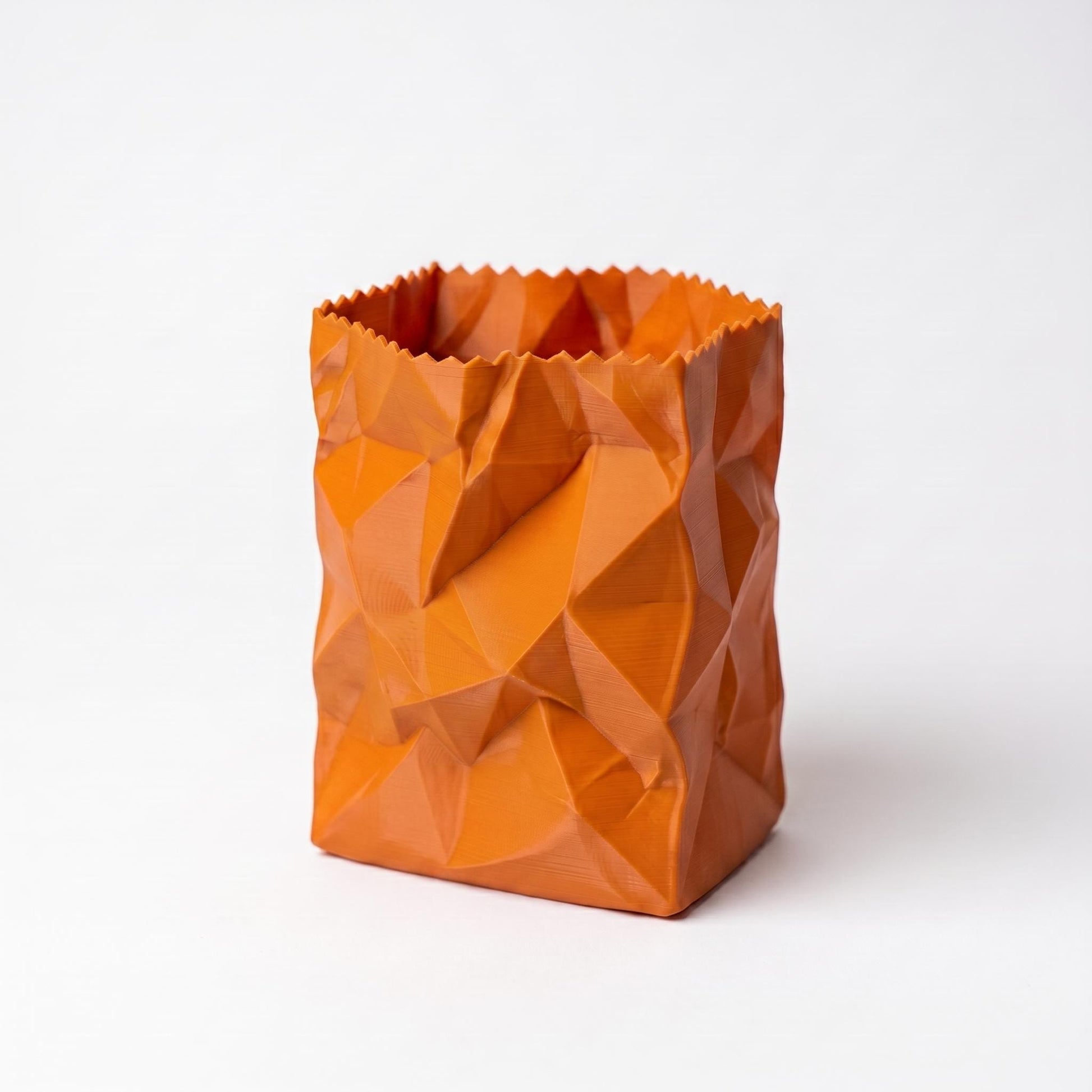 Ein orangefarbener 3D-gedruckter Stiftehalter in zerknitterter Papiertüten-Optik steht auf weißem Hintergrund. Das geometrische Design zeigt facettierte Oberflächen mit Dreiecksmustern und einem gezackten oberen Rand. Der pen holder aus PLA-Filament eignet sich perfekt als desk organizer und moderne Bürodeko. Ideal als Schreibtisch Organizer für Stifte und Büroartikel.