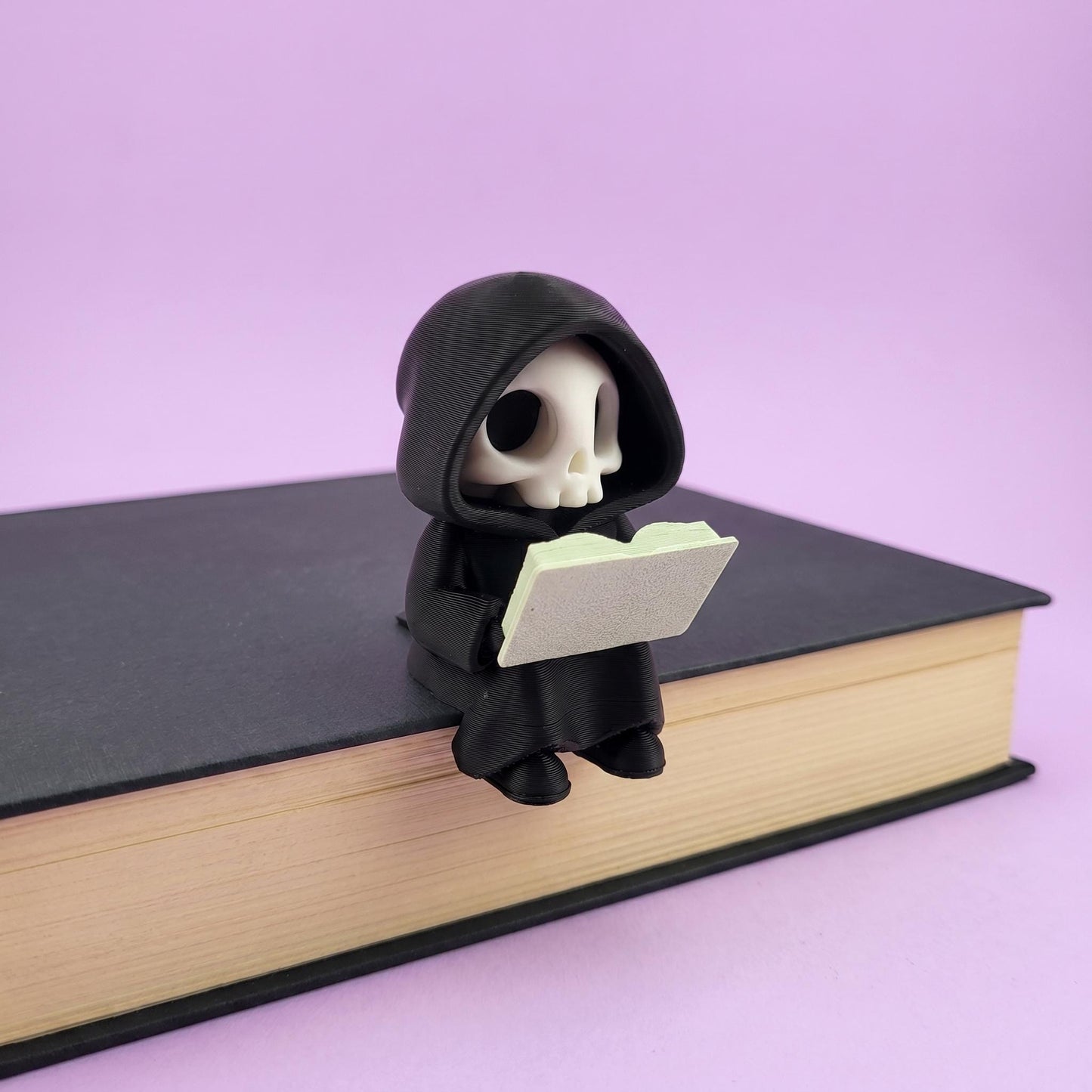 3D-gedruckte Sensenmann-Figur sitzt lesend auf einem Buch, schwarzer Kapuzenumhang mit weißem Totenkopf, vor hellem fliederfarbenem Hintergrund