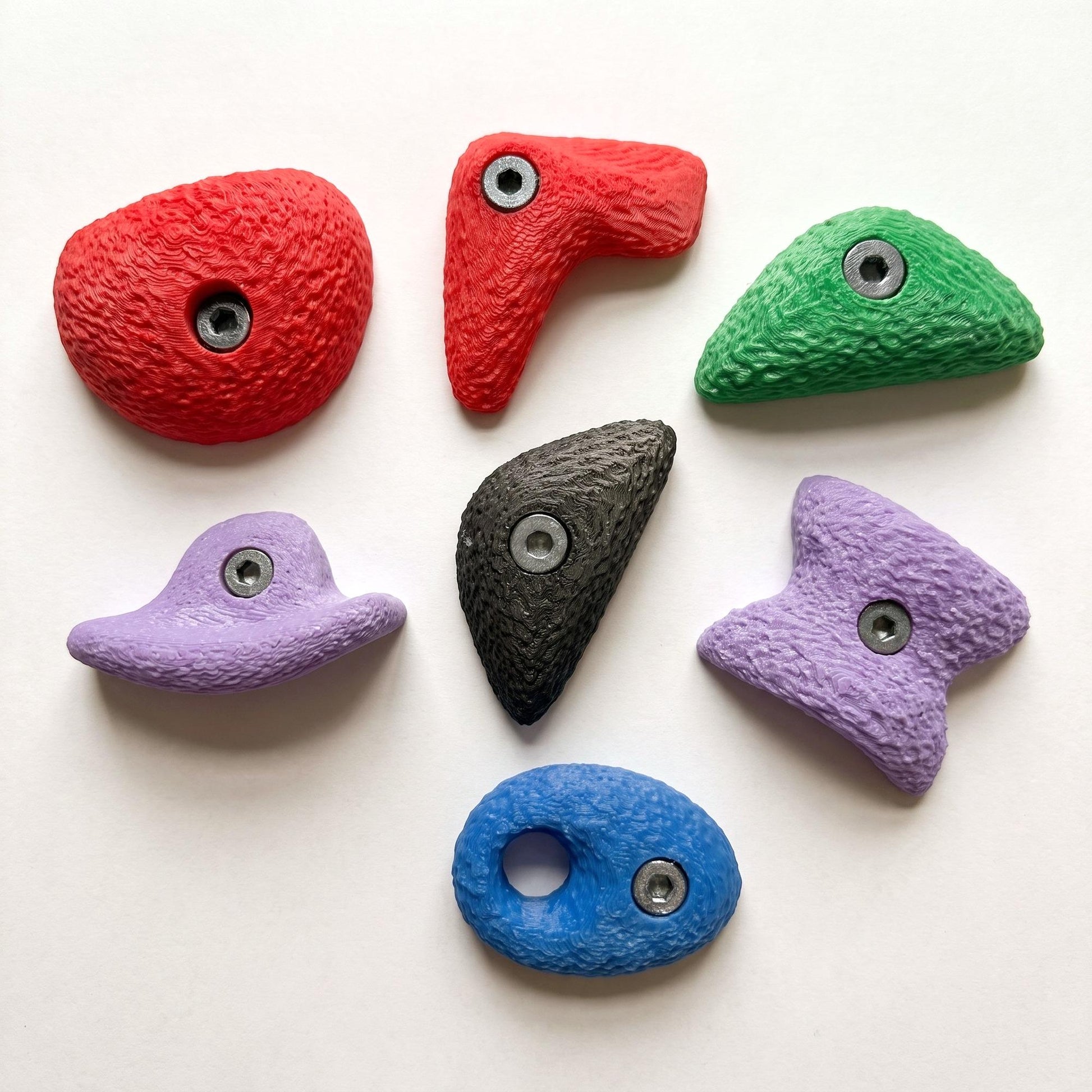 Alle sieben großen Bouldergriff-Magnete im Farbmix, locker angeordnet auf hellem Grund — Kühlschrankmagnete in echten Klettergriff-Formen, handgefertigt im 3D-Druck.