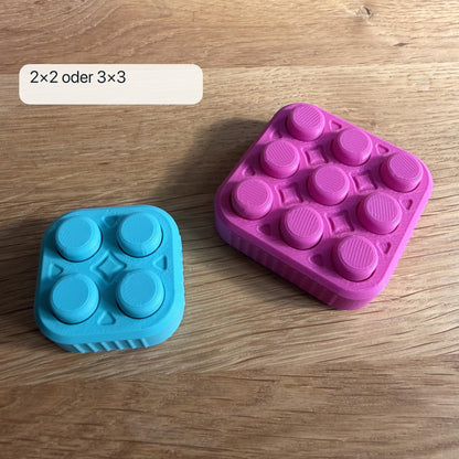 Direkter Größenvergleich zwischen einem türkisen 2×2 Bubble Popper mit vier und einem magentafarbenen 3×3 Popper mit neun Druckflächen.