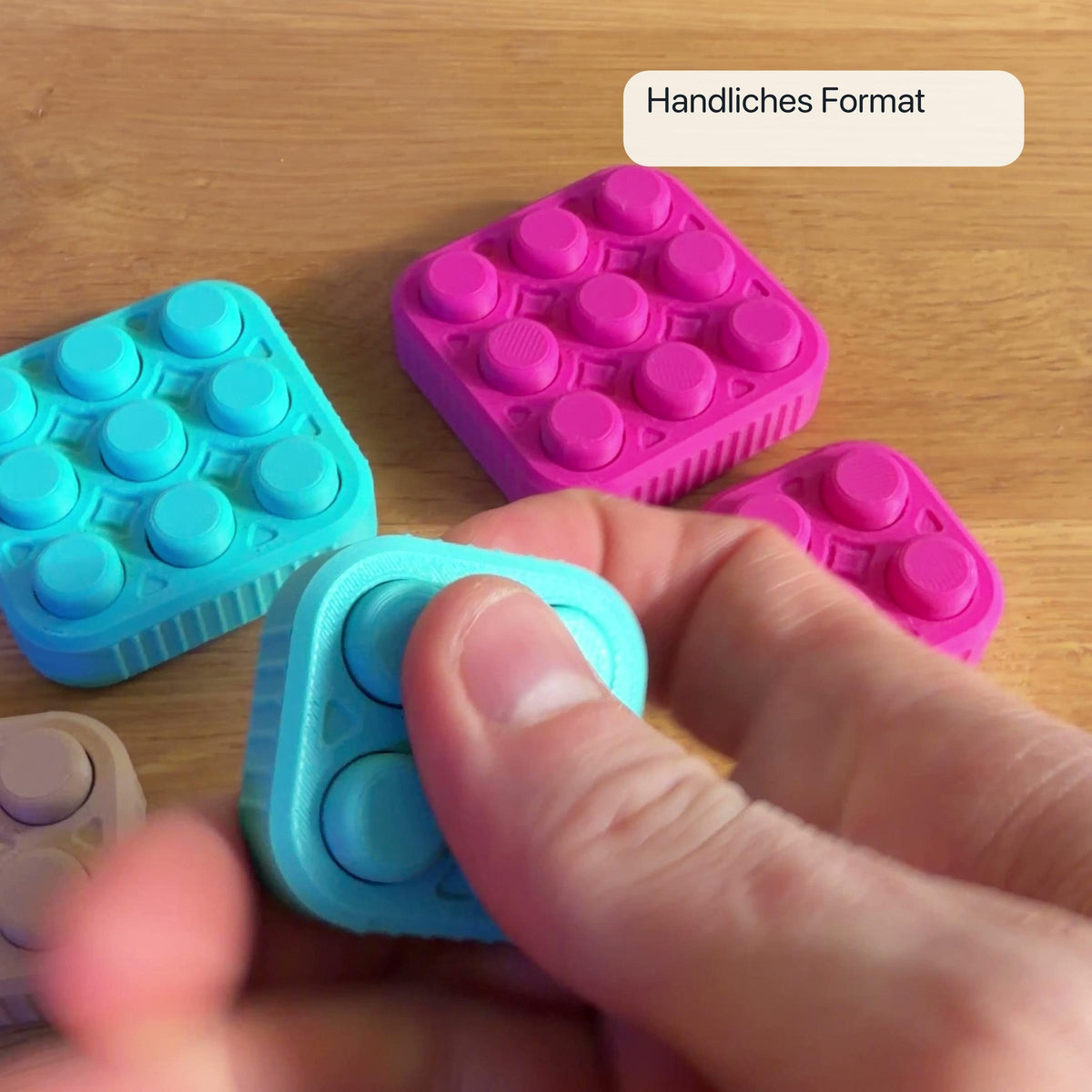 Ein türkiser 2×2 Bubble Popper wird in einer Hand gehalten; weitere Popper in Türkis und Magenta liegen im Hintergrund.