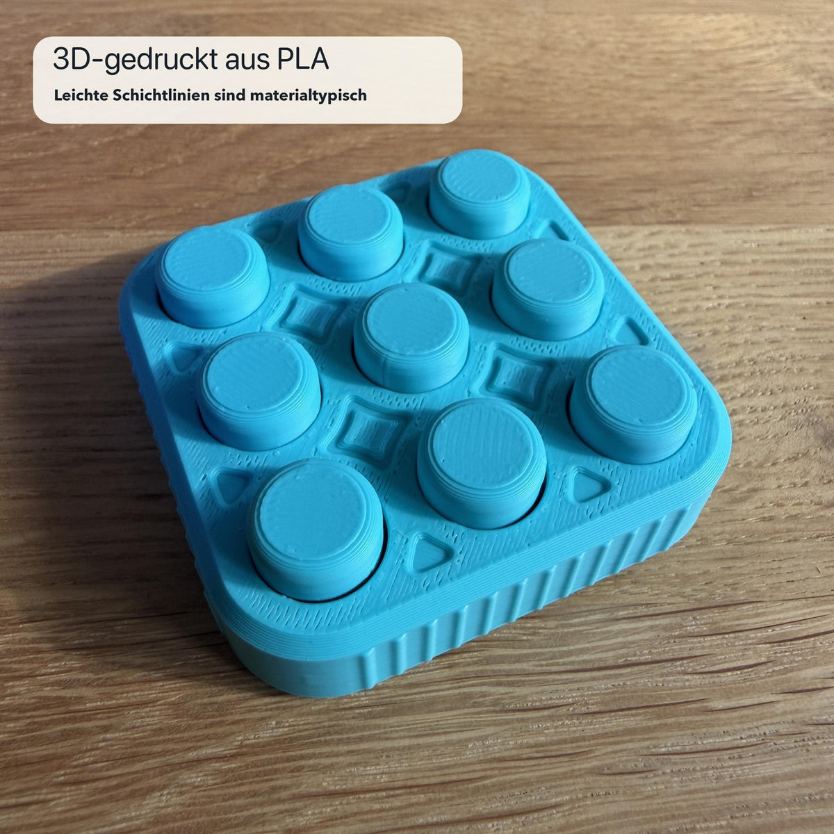 Türkiser 3×3 Bubble Popper aus PLA, schräg von oben auf einem Holztisch fotografiert; Schichtlinien und geriffelte Seitenstruktur des 3D-Drucks sind erkennbar.