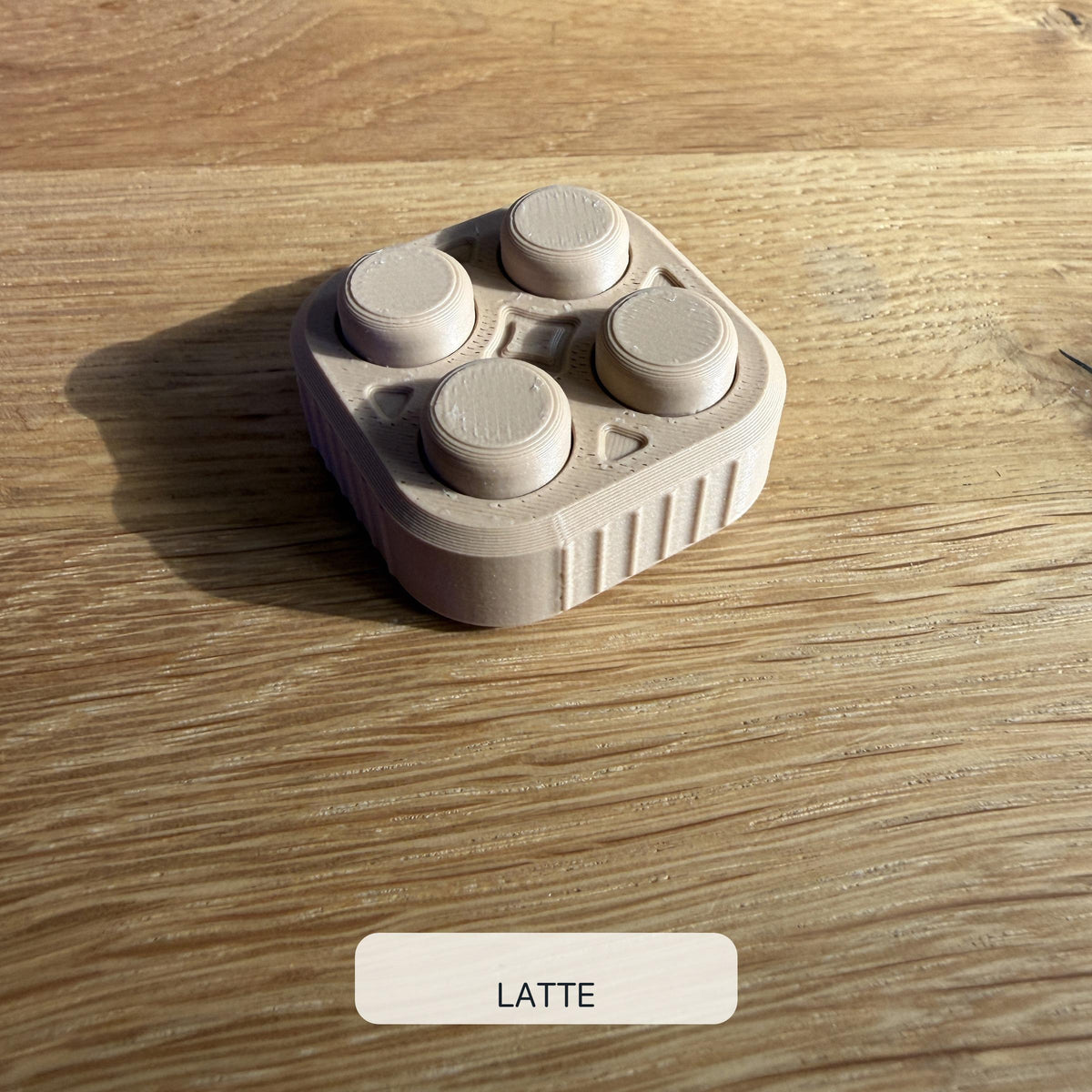 Dezent lattebrauner beziehungsweise beigefarbener 2×2 Bubble Popper mit vier runden Druckflächen, schräg von oben auf einem Holztisch fotografiert.