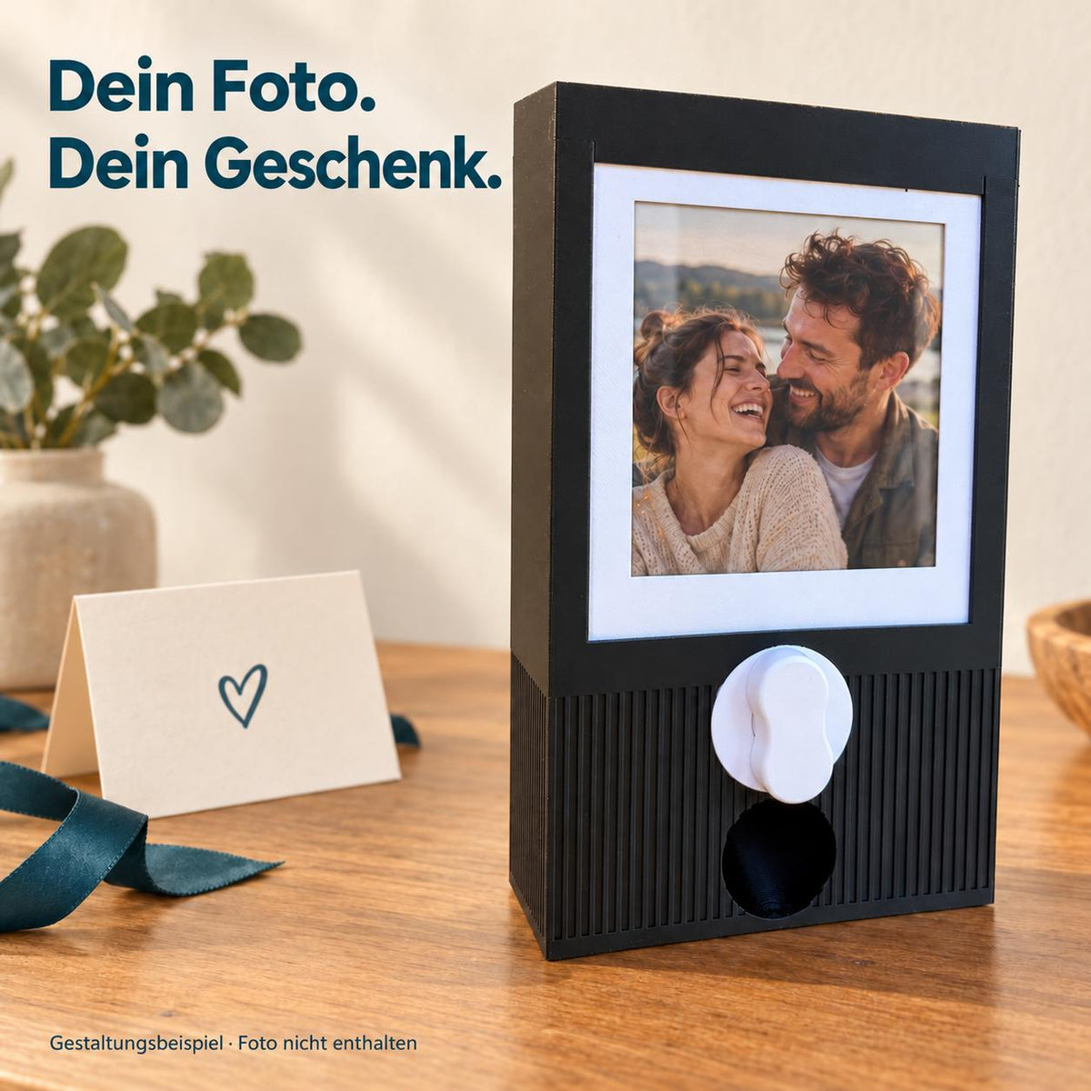 Schwarzer Kapselautomat mit einem beispielhaften gedruckten Paarfoto im weißen Einsteckrahmen. Überschrift: Dein Foto. Dein Geschenk. Foto und Dekoration mit KI ergänzt und nicht enthalten; Fotos werden vom Käufer selbst eingesetzt.