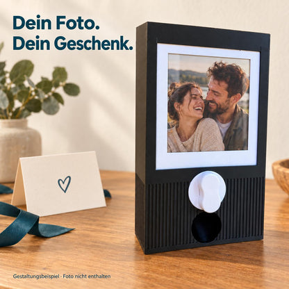 Schwarzer Kapselautomat mit einem beispielhaften gedruckten Paarfoto im weißen Einsteckrahmen. Überschrift: Dein Foto. Dein Geschenk. Foto und Dekoration mit KI ergänzt und nicht enthalten; Fotos werden vom Käufer selbst eingesetzt.