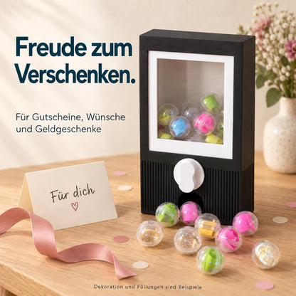 Freude zum Verschenken. Schwarzer Kapselautomat für Gutscheine, Wünsche und Geldgeschenke. KI-gestaltetes Dekobeispiel mit Karte und Blüten; Dekoration und gezeigte Füllungen sind nicht enthalten.